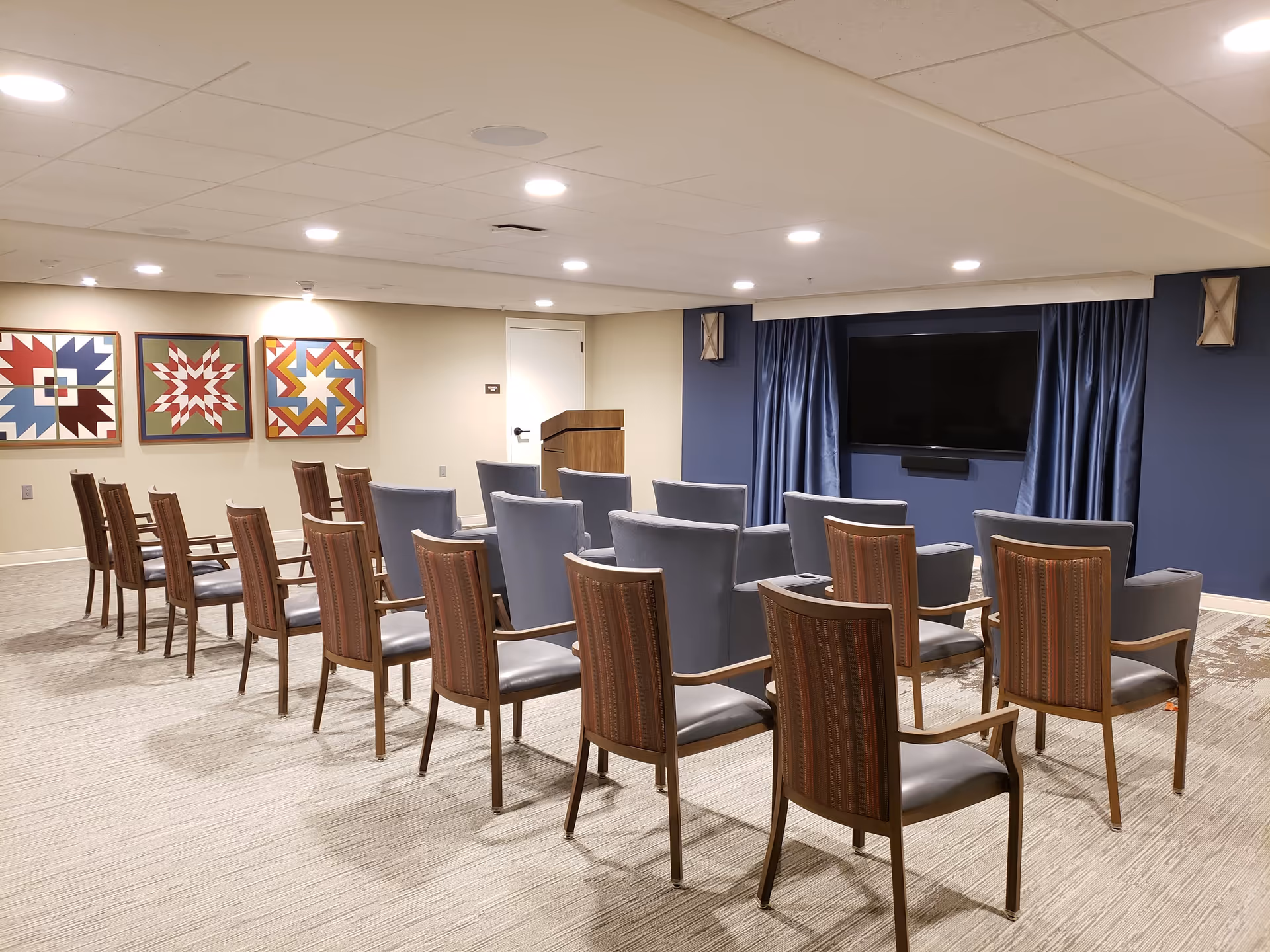 A small auditorium or meeting room with rows of chairs facing a large flat-screen TV mounted on a blue accent wall with blue curtains on either side. There is a wooden podium near a white door on the back wall, and three colorful framed quilt-like artworks hang on the opposite wall. The room is well-lit with recessed ceiling lights.