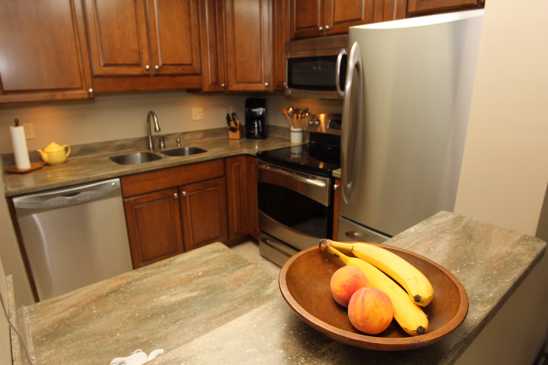 Small kitchen with wooden cabinets, stainless steel appliances, a double sink and a bowl of bananas and peaches on the counter.