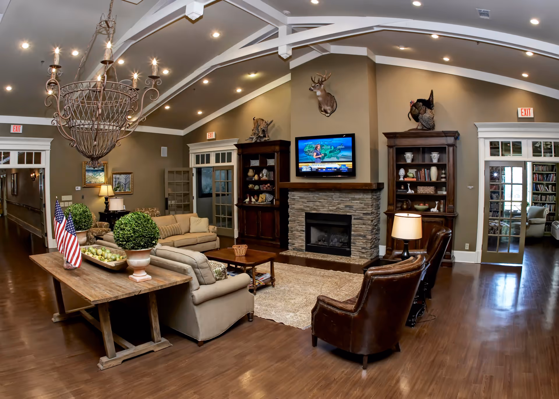 A cozy living room in an assisted living community featuring a stone fireplace with a mounted deer head above it, a flat-screen TV displaying a weather forecast, two wooden bookshelves filled with decorative items, a beige sofa and loveseat, two brown leather armchairs, a wooden coffee table, and a rustic wooden table with two potted plants and two small American flags. The room has wooden flooring, recessed lighting, and a large decorative chandelier hanging from a vaulted ceiling. French doors lead to another room with bookshelves and seating.