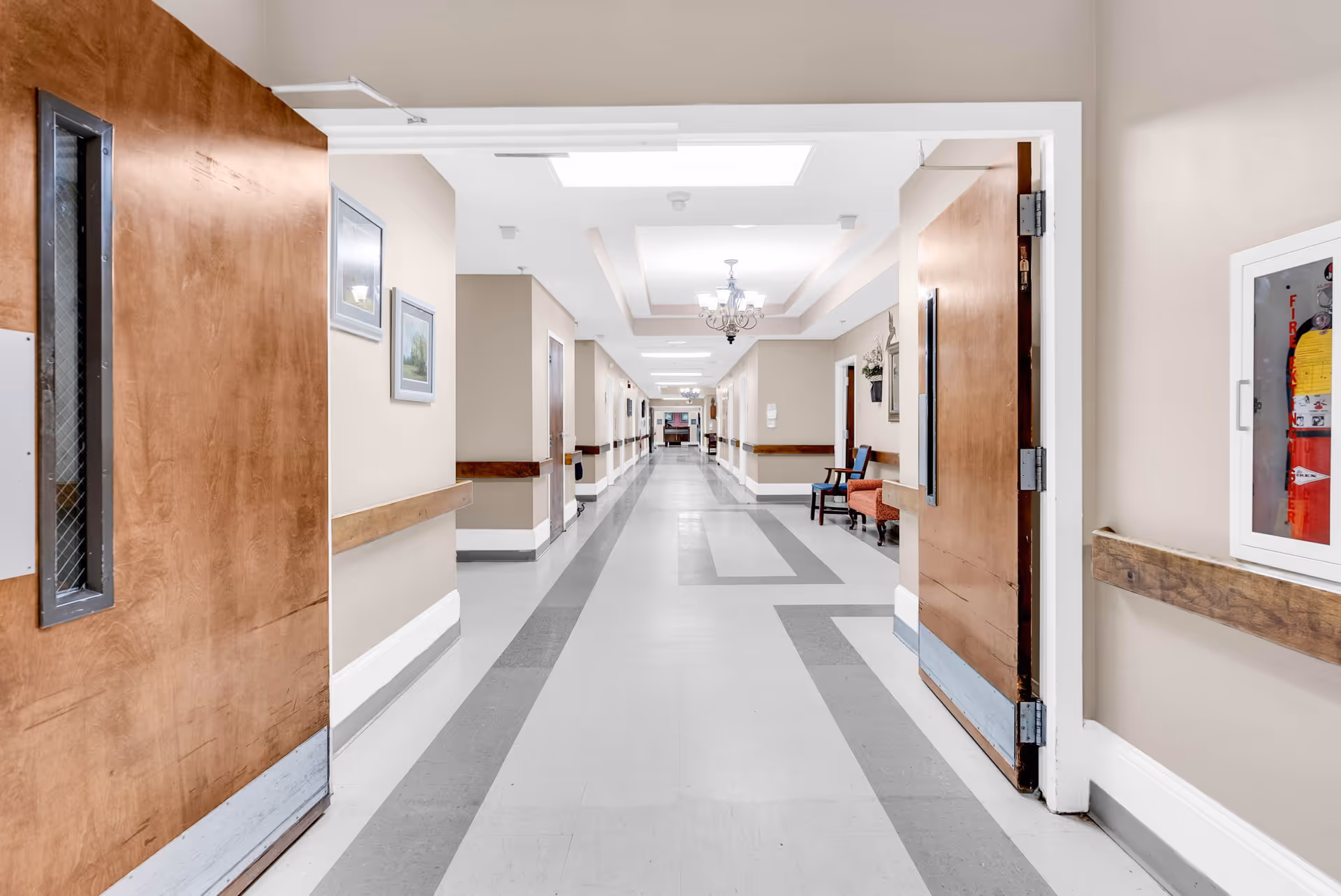 A long, clean hallway in a healthcare facility with beige walls, gray and white patterned flooring, wooden handrails, and open wooden doors on either side. The hallway is well-lit with ceiling lights and chandeliers, and there are chairs and framed pictures along the walls.