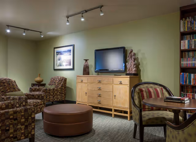 Cozy lounge with patterned armchairs, a round ottoman, a wooden media cabinet with a TV, and a bookshelf.