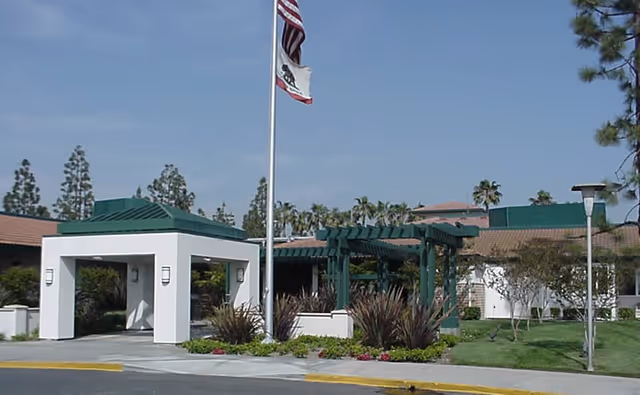 Front entrance of a senior living facility showing a porte-cochère, flagpoles, a green pergola, and landscaped grounds.