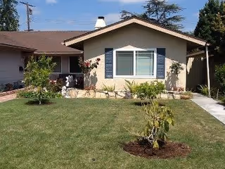 Single-story house with beige exterior walls, a brown roof, and blue window shutters. The front yard has a well-maintained green lawn with small trees and plants. A concrete pathway leads to the entrance on the right side of the house.