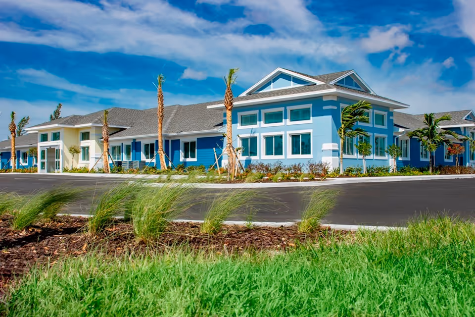 Exterior view of a single-story senior living facility building with blue and white walls, multiple windows, palm trees, and a clear blue sky with some clouds.