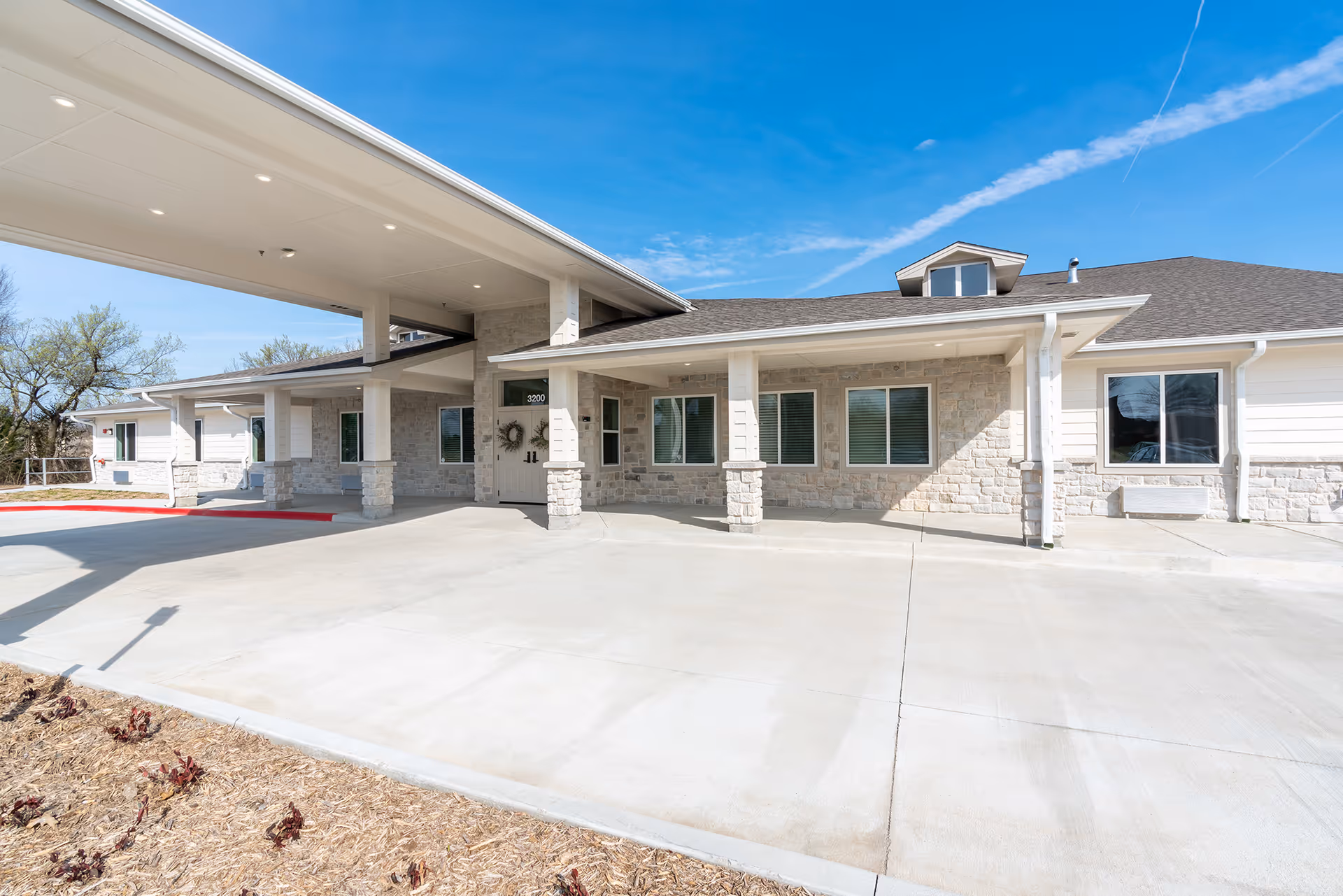 Front exterior view of BeeHive Homes of Broken Arrow building with a covered entrance, stone and siding facade, several windows, and a clear blue sky above.