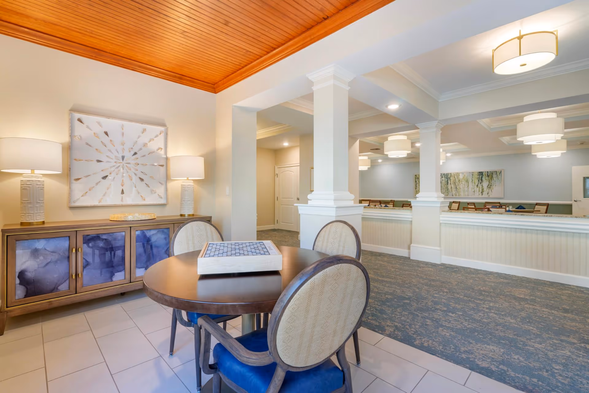 A well-lit interior space featuring a round table with four upholstered chairs, a decorative sideboard with two lamps and artwork above it, and a carpeted area with columns and ceiling lights in the background.