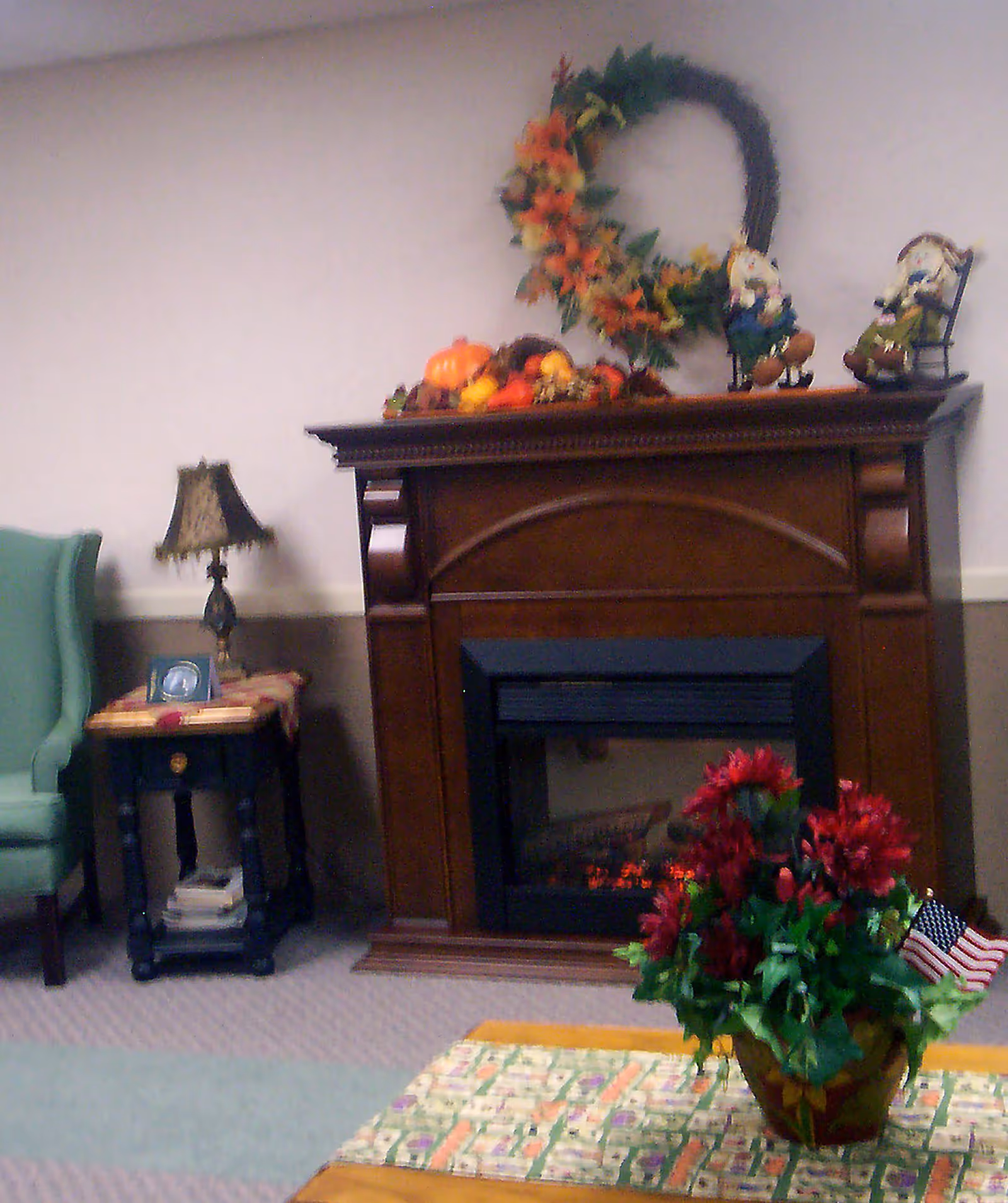 A cozy living room area featuring a wooden electric fireplace decorated with a fall-themed wreath and small figurines on the mantel. To the left is a green upholstered armchair and a small side table with a decorative lamp and framed photo. In the foreground, there is a table with a floral arrangement and a small American flag.