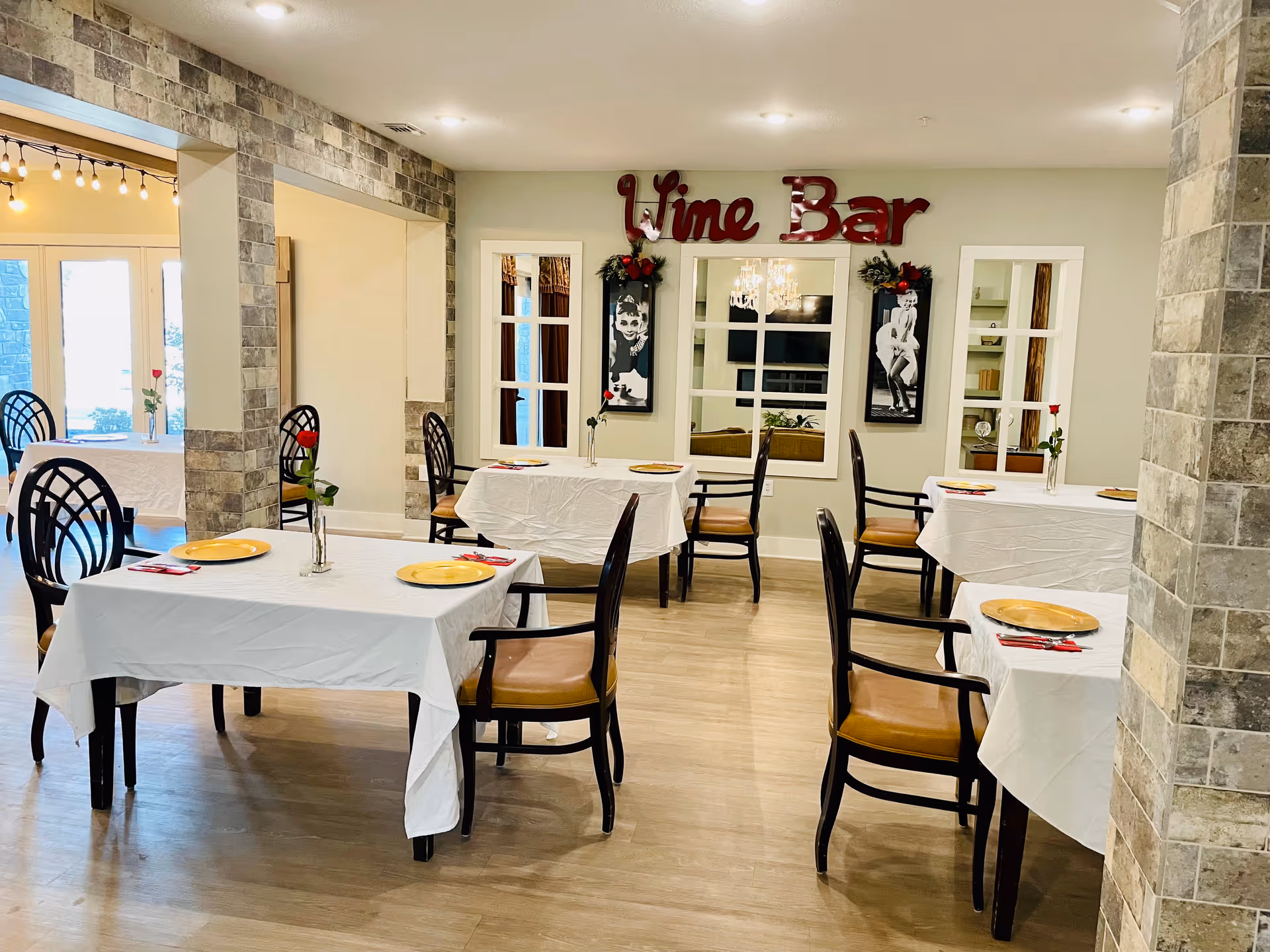 A dining area with several tables covered in white tablecloths, each set with a yellow plate, red napkin, and a single red rose in a vase. The room has wooden flooring and stone pillars. On the far wall, there is a decorative sign that reads 'Wine Bar' above two black and white framed pictures and a window reflecting a chandelier and TV.