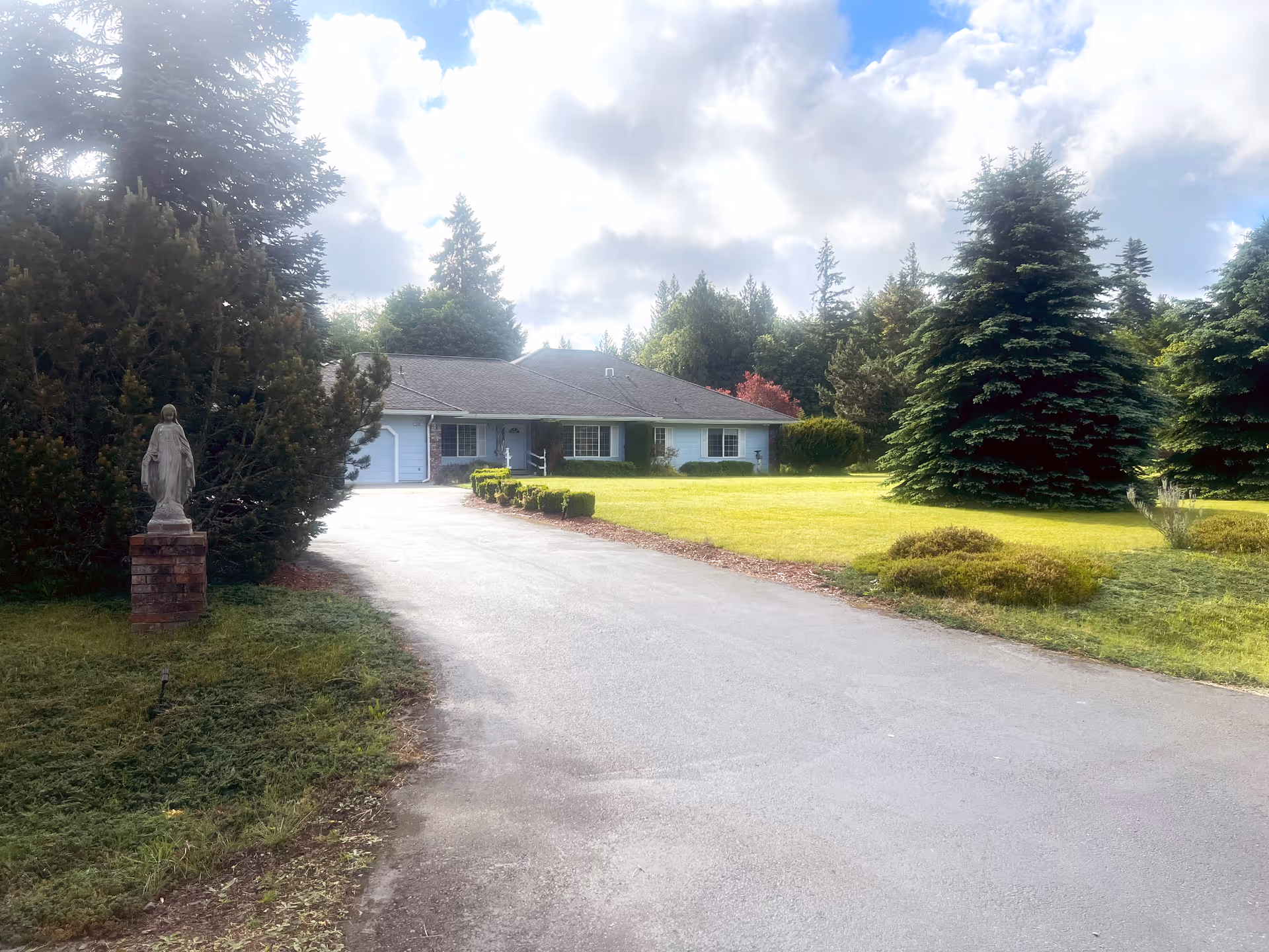 A single-story house with a gray roof and white exterior surrounded by a large, well-maintained lawn and tall evergreen trees under a partly cloudy sky. A paved driveway leads up to the house, and there is a statue on a brick pedestal near the driveway on the left side.