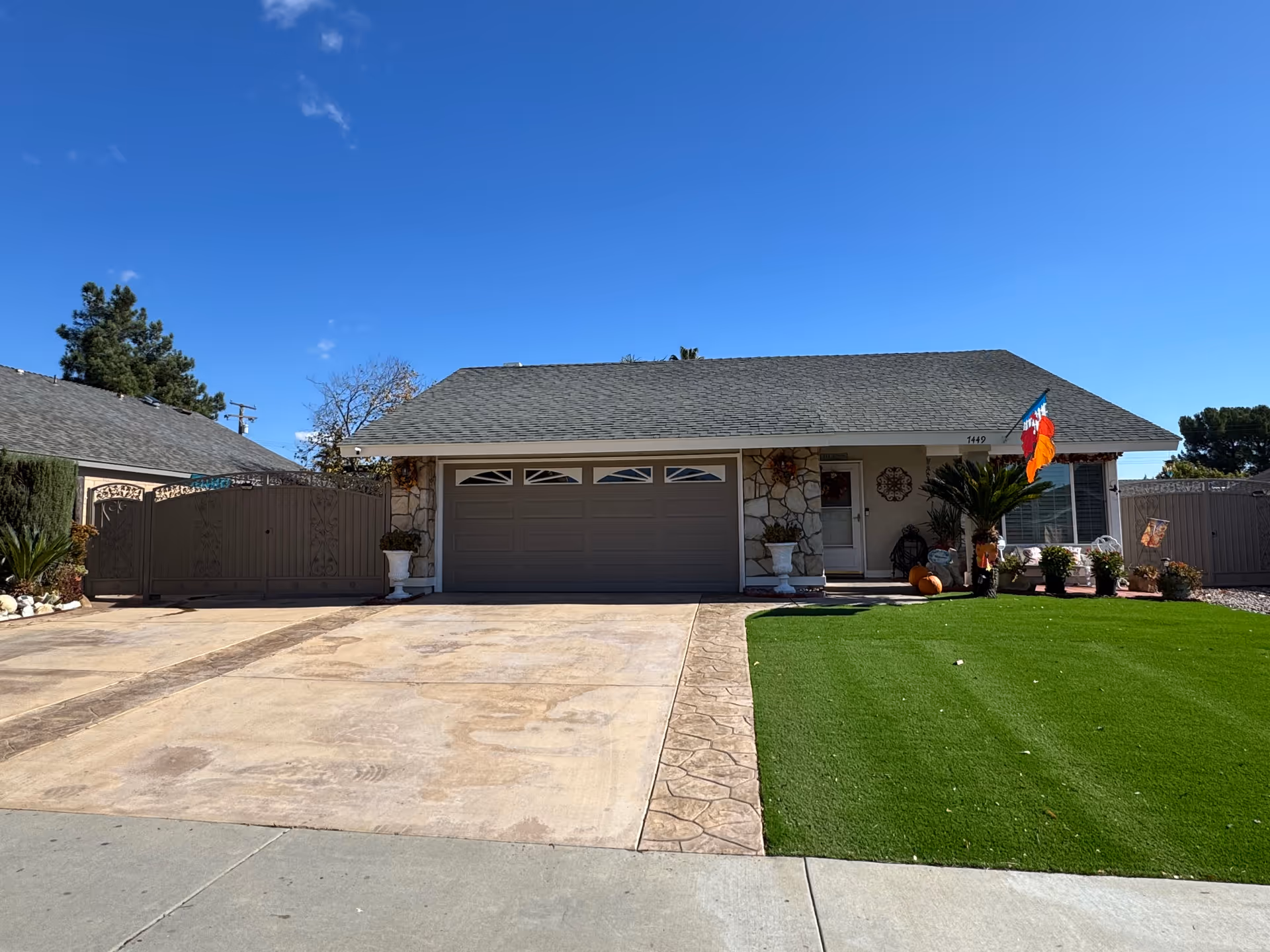 Single-story house with a gray roof and stone facade, featuring a double garage door and a well-maintained front yard with green artificial grass. There are potted plants and a flag near the entrance, under a clear blue sky.
