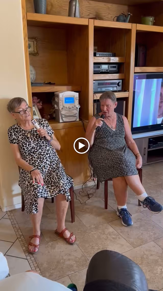 Two elderly women sitting on chairs in a living room, each holding a microphone and appearing to sing karaoke. Behind them is a wooden entertainment center with audio equipment and a large TV screen.