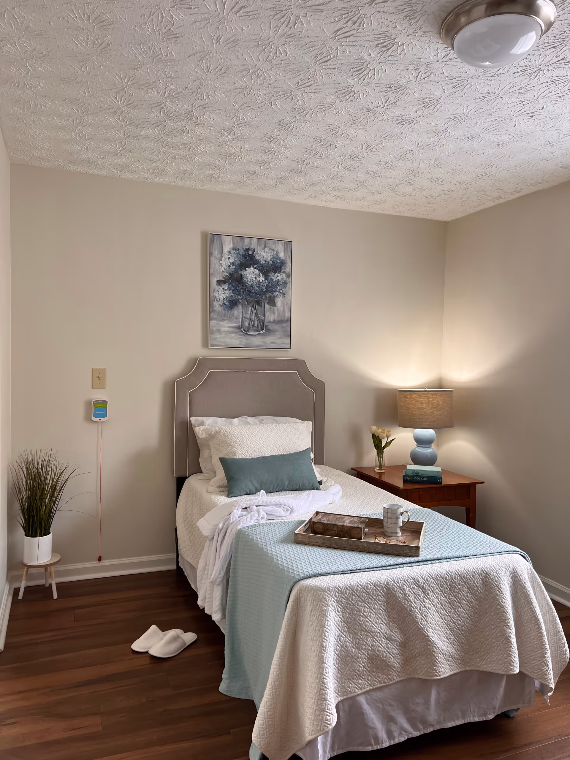A neatly made single bed with white and light blue bedding in a cozy bedroom. There is a small wooden nightstand with a blue lamp, a vase with flowers, and two books. A painting of blue and white flowers hangs on the beige wall above the bed. A pair of white slippers is placed on the wooden floor next to the bed, and a small potted plant stands on a stool in the corner. The ceiling has a textured pattern with a round light fixture.