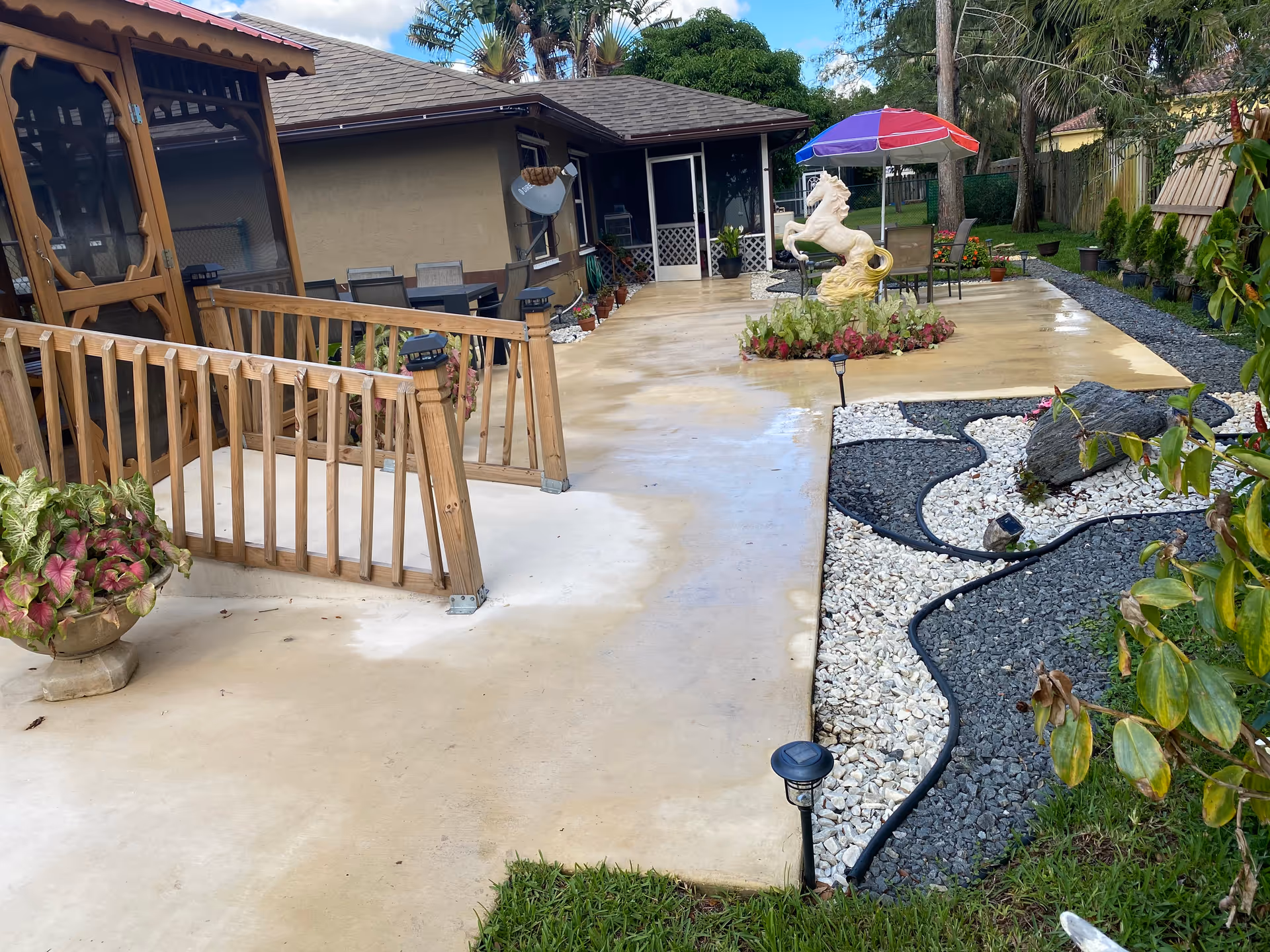 Outdoor patio area of a senior living facility with a concrete walkway, a wooden railing, a gazebo, a table with chairs under a colorful umbrella, a white horse statue surrounded by plants, and decorative landscaping with white and black rocks.