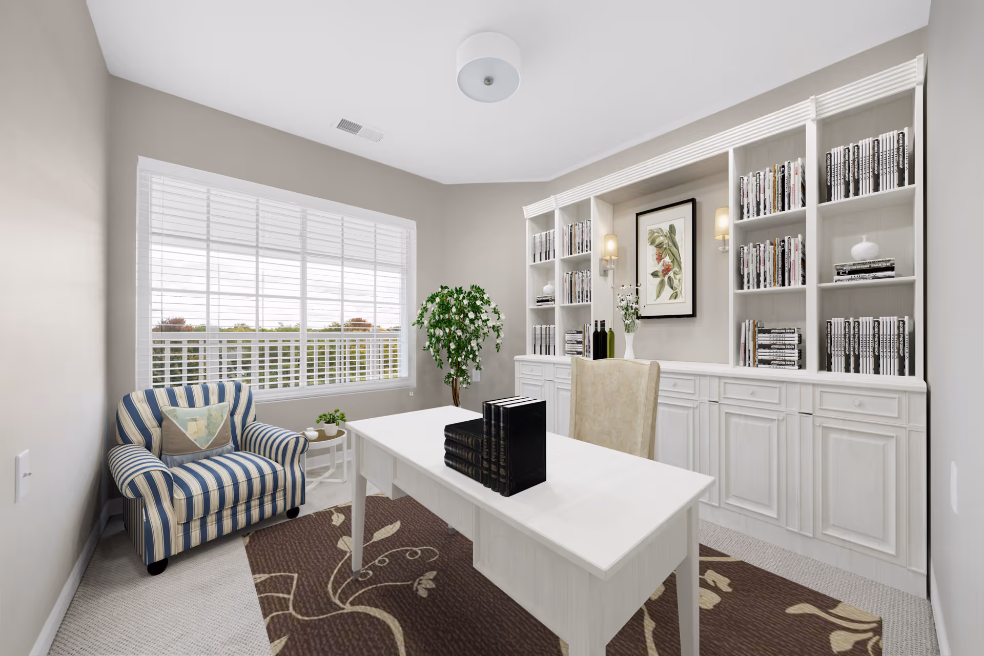 A bright office or study room with a large window featuring white blinds. The room has a white desk with a set of black books on it, a beige upholstered chair, and a striped armchair with a decorative pillow. There is a large white built-in bookshelf filled with books and decorative items, a framed botanical print on the wall, and a potted plant in the corner. The floor has a patterned brown rug.