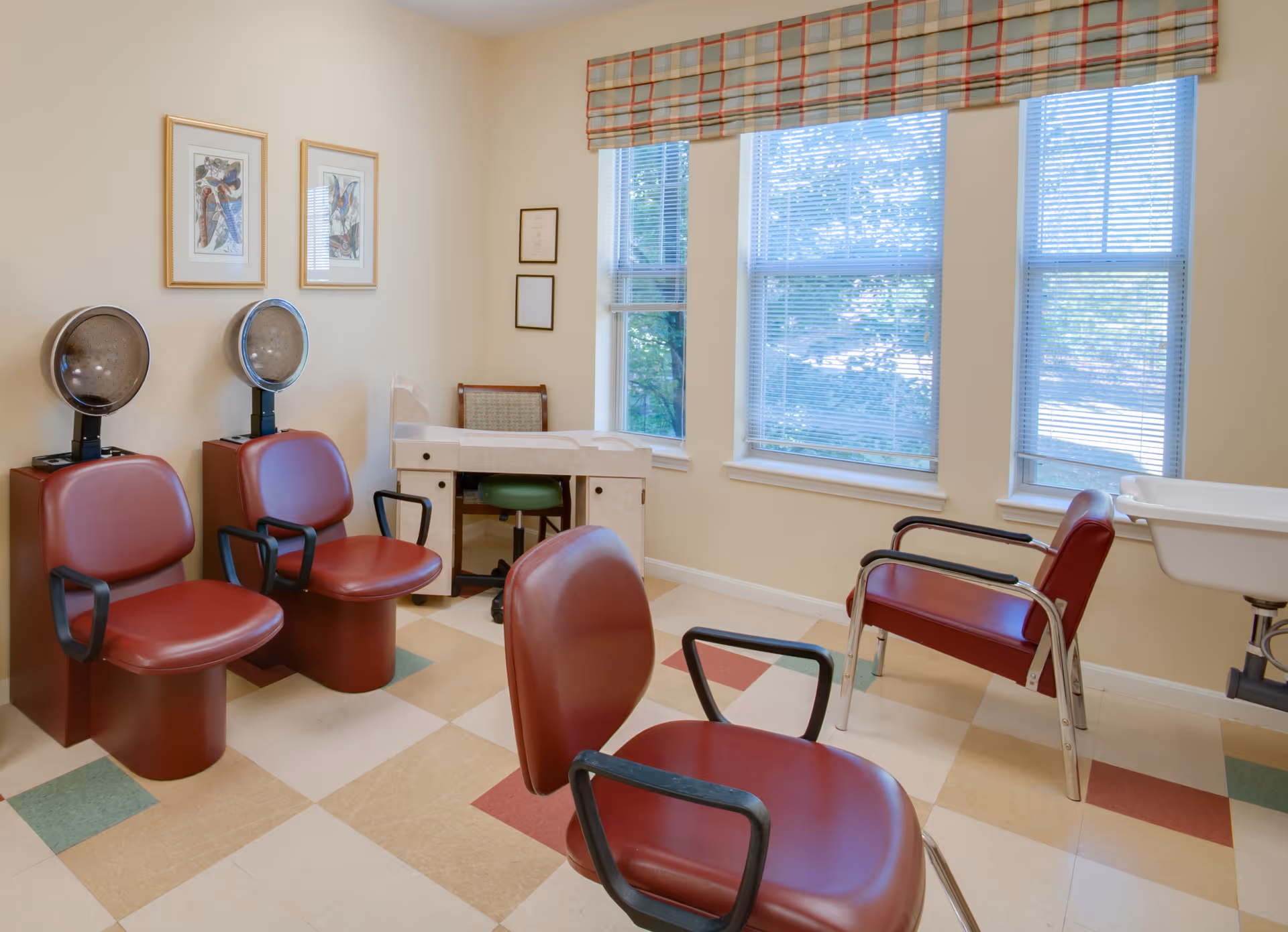 A small salon or hair care room with four red salon chairs, two with attached hair dryers. The room has a checkered floor with beige, red, and green tiles, three large windows with blinds and a plaid valance, two framed pictures on the wall, and a small desk with a green chair in the corner.