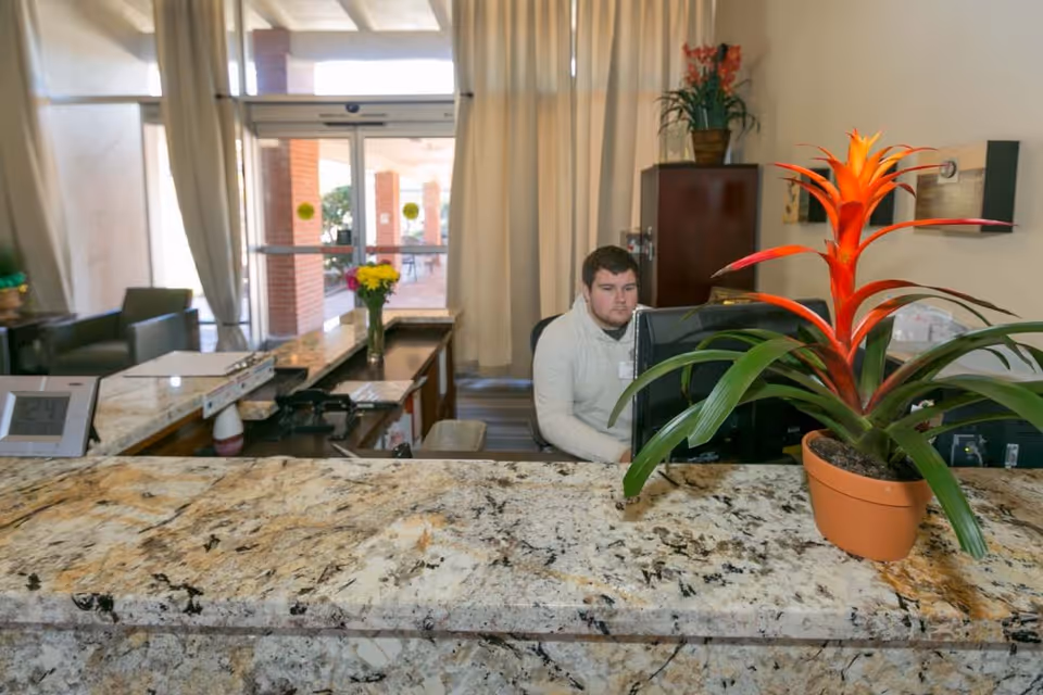 Reception area with a man sitting behind a granite countertop desk, working on a computer. There is a potted plant with bright orange flowers on the counter, a vase with yellow flowers, and a cabinet with another plant in the background. Glass doors lead to an outdoor area.