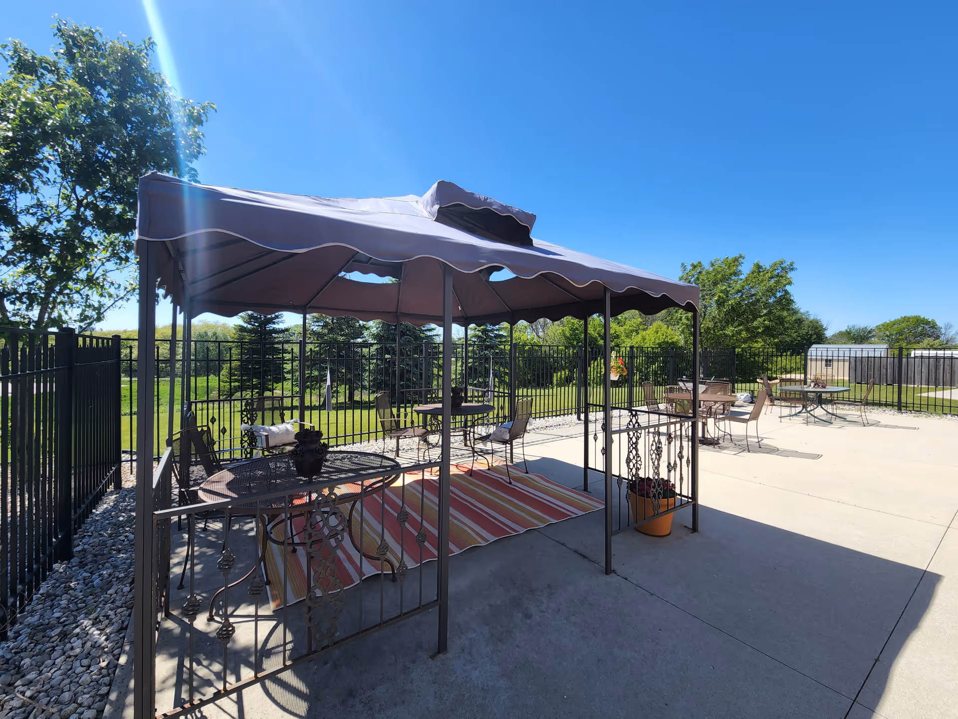Sunlit outdoor patio with a canopied gazebo, metal tables and chairs, a striped rug, and a fenced green lawn with trees.