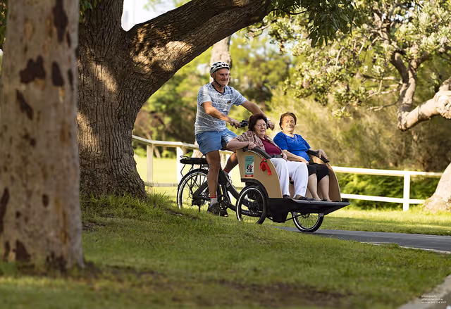 A man wearing a helmet rides a bicycle with a front passenger seat carrying two elderly women along a paved path in a park-like setting with trees and grass.