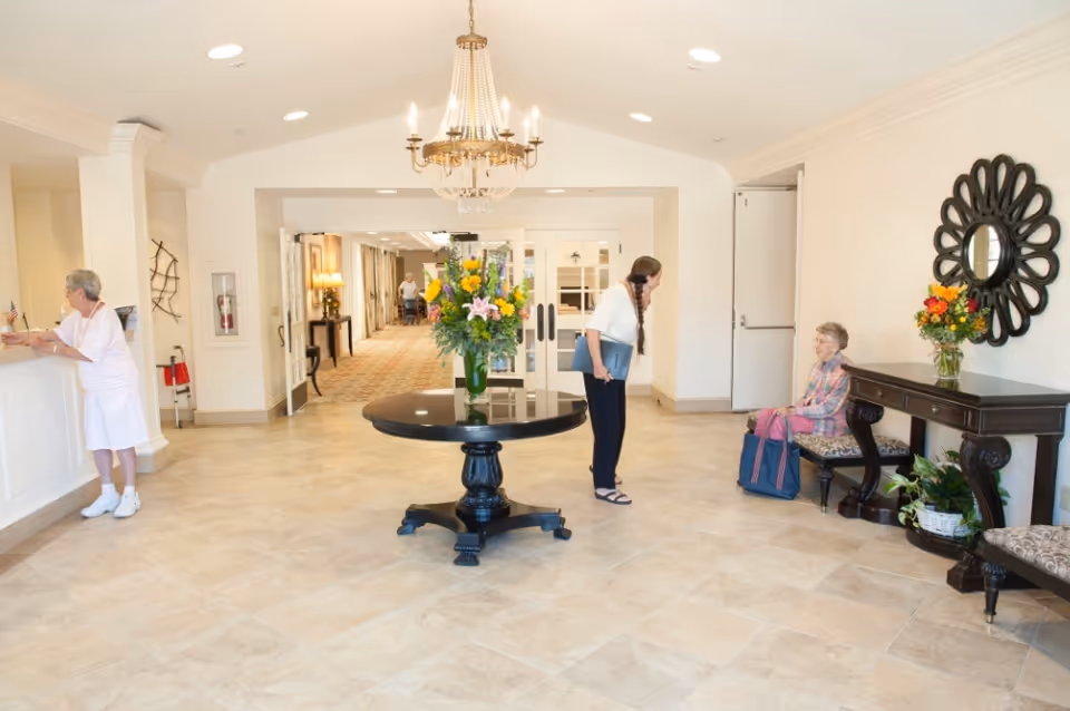 Bright senior living lobby with a round table topped with flowers, chandelier, reception desk and residents interacting.
