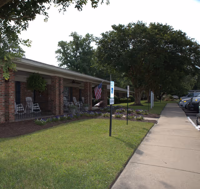 Exterior view of a single-story brick building with a covered porch featuring several rocking chairs and hanging plants. An American flag is displayed near the entrance. There is a well-maintained lawn with flower beds and a sidewalk leading to a parking area with several cars parked. Trees provide shade along the sidewalk.