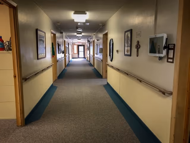 A long, well-lit hallway in a senior living facility with beige walls and blue trim. The hallway has handrails on both sides and several doors leading to rooms. There are framed pictures and a cross hanging on the walls, and a tablet device mounted near one of the doors labeled 102. The hallway extends to a glass exit door at the far end.