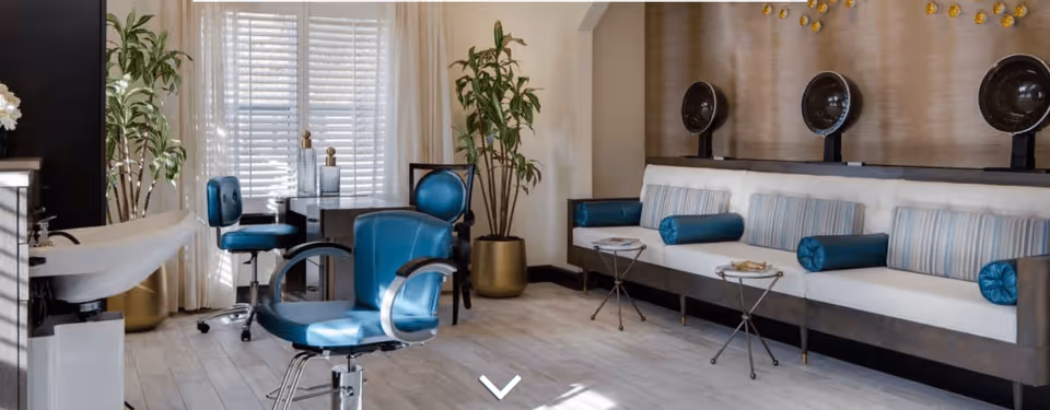 A stylish salon area with blue salon chairs, a white sink, and a long white cushioned bench with blue and striped pillows. There are three vintage hair dryers mounted on the wall above the bench, potted plants, and a window with white shutters and sheer curtains letting in natural light.