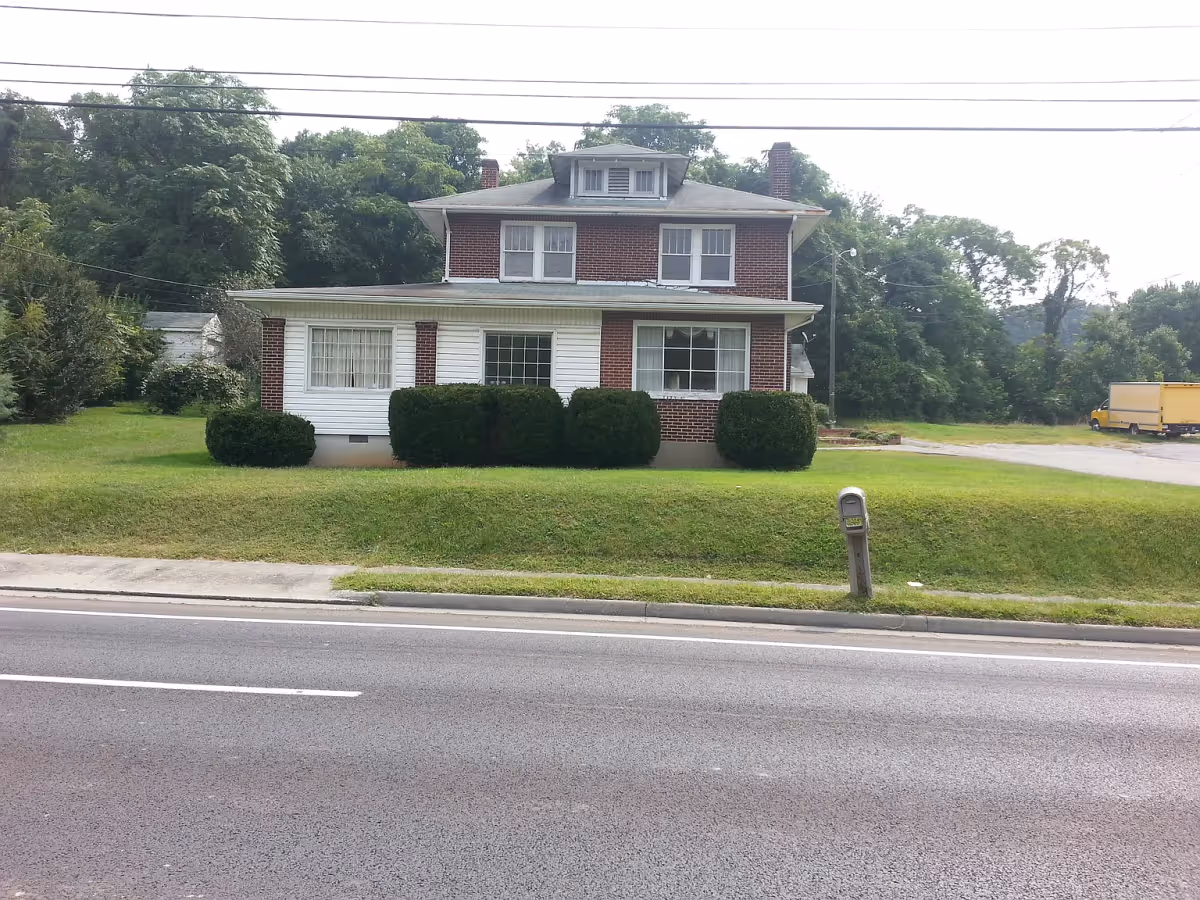 Front exterior view of a two-story brick and white siding house with several windows, surrounded by green bushes and grass, with a mailbox near the road and trees in the background.