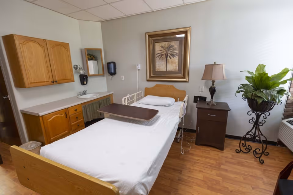 A clean and simple patient room in a care facility featuring a single hospital bed with white linens and a wooden headboard. The bed has a movable overbed table. To the left, there is a countertop with a sink, wooden cabinets above and below, a mirror, and a soap dispenser. On the right side of the bed, there is a dark wooden nightstand with a lamp and a large potted plant on a decorative metal stand near a window. A framed picture of a palm tree hangs on the wall above the bed.