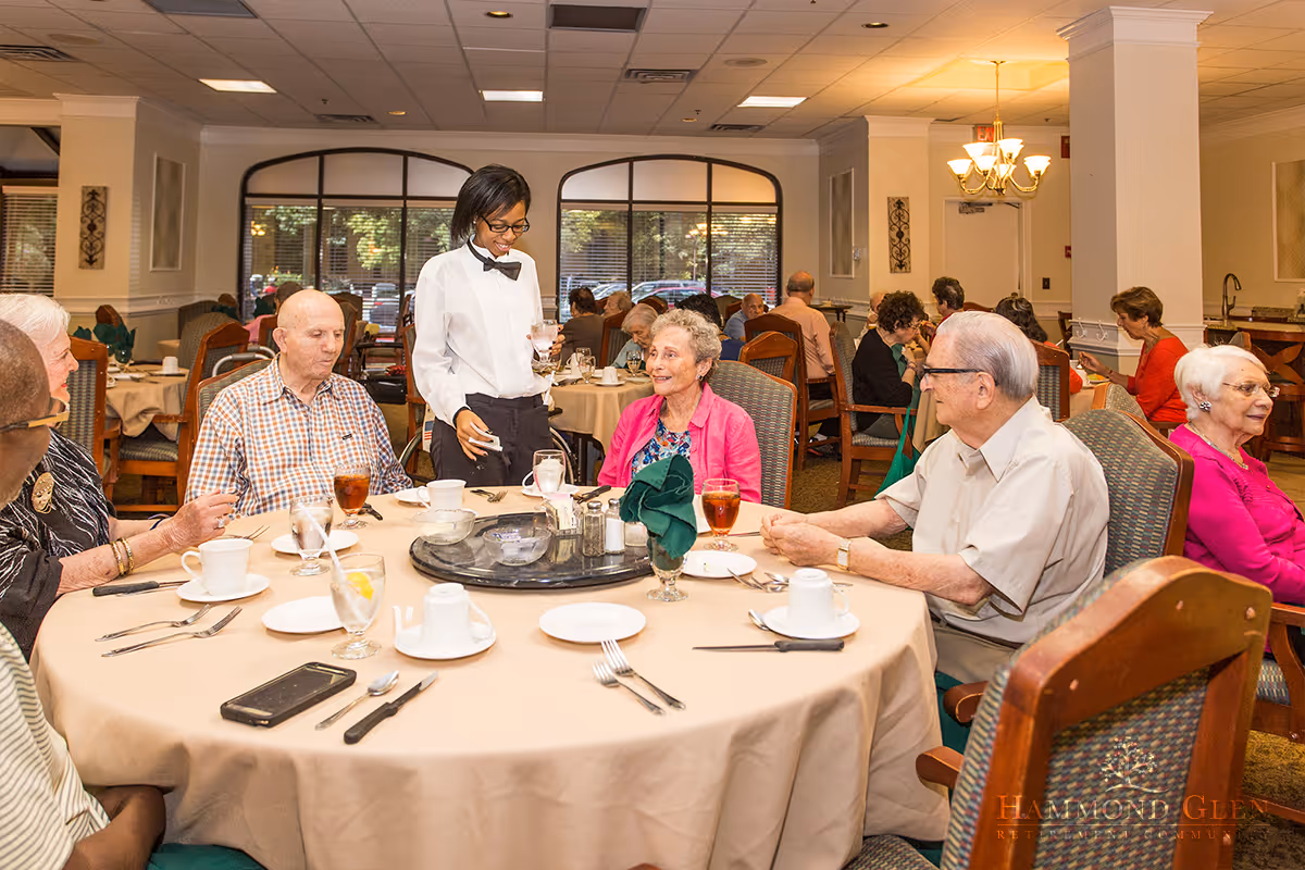 A group of elderly people sitting around a round dining table in a retirement community dining room. A waitress in a white shirt and black bow tie is serving water to one of the residents. The table is set with cups, glasses, plates, and utensils. Other residents are seated at tables in the background, and large windows let in natural light.