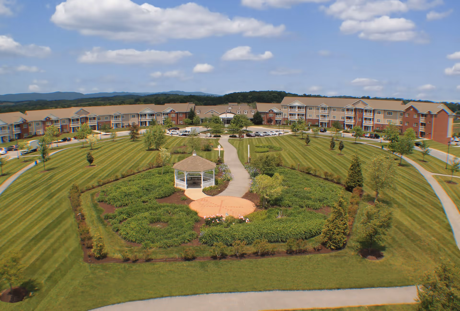 Aerial view of The Glebe Retirement Community showing a large, well-maintained garden with a white gazebo in the center, surrounded by green lawns, trees, and shrubs. In the background, there is a multi-story residential building with balconies and a parking area with several cars. The sky is partly cloudy with blue patches.