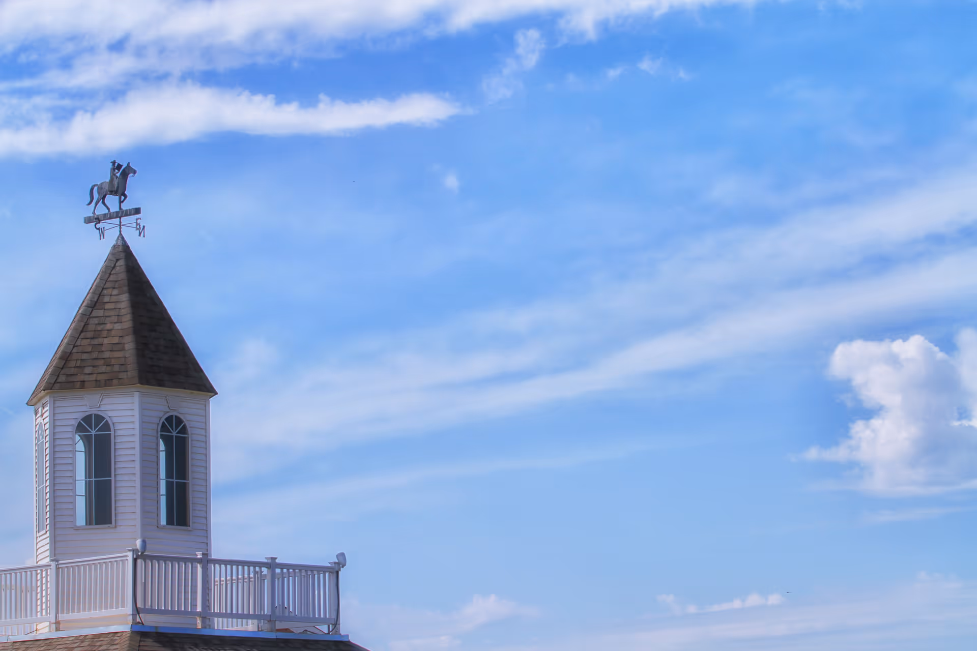 A white cupola with arched windows and a shingled roof topped by a weather vane featuring a horse and rider, set against a blue sky with scattered clouds.
