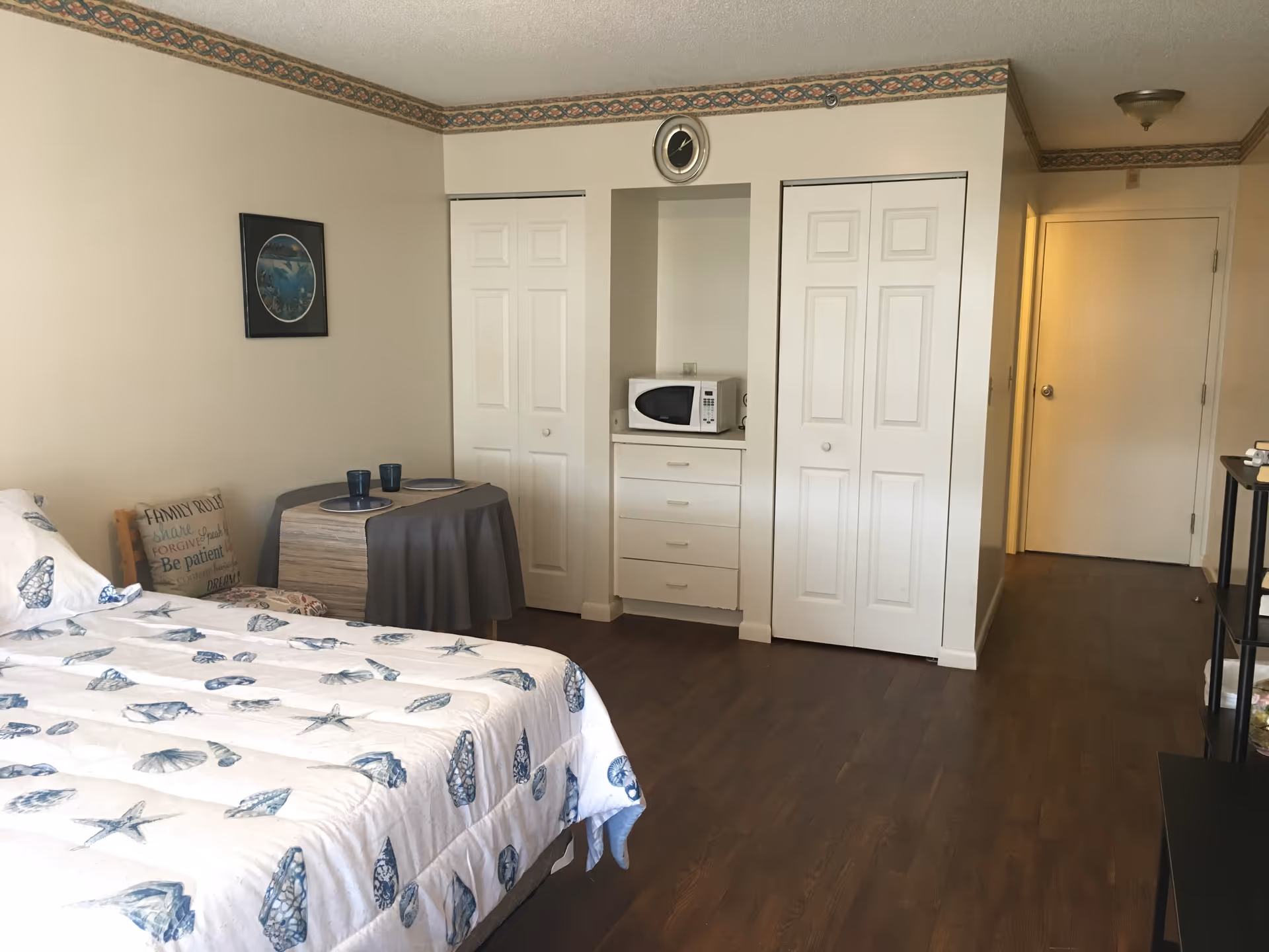Studio-style bedroom with a bed, small table, built-in closets and a microwave on drawers, and wood flooring.