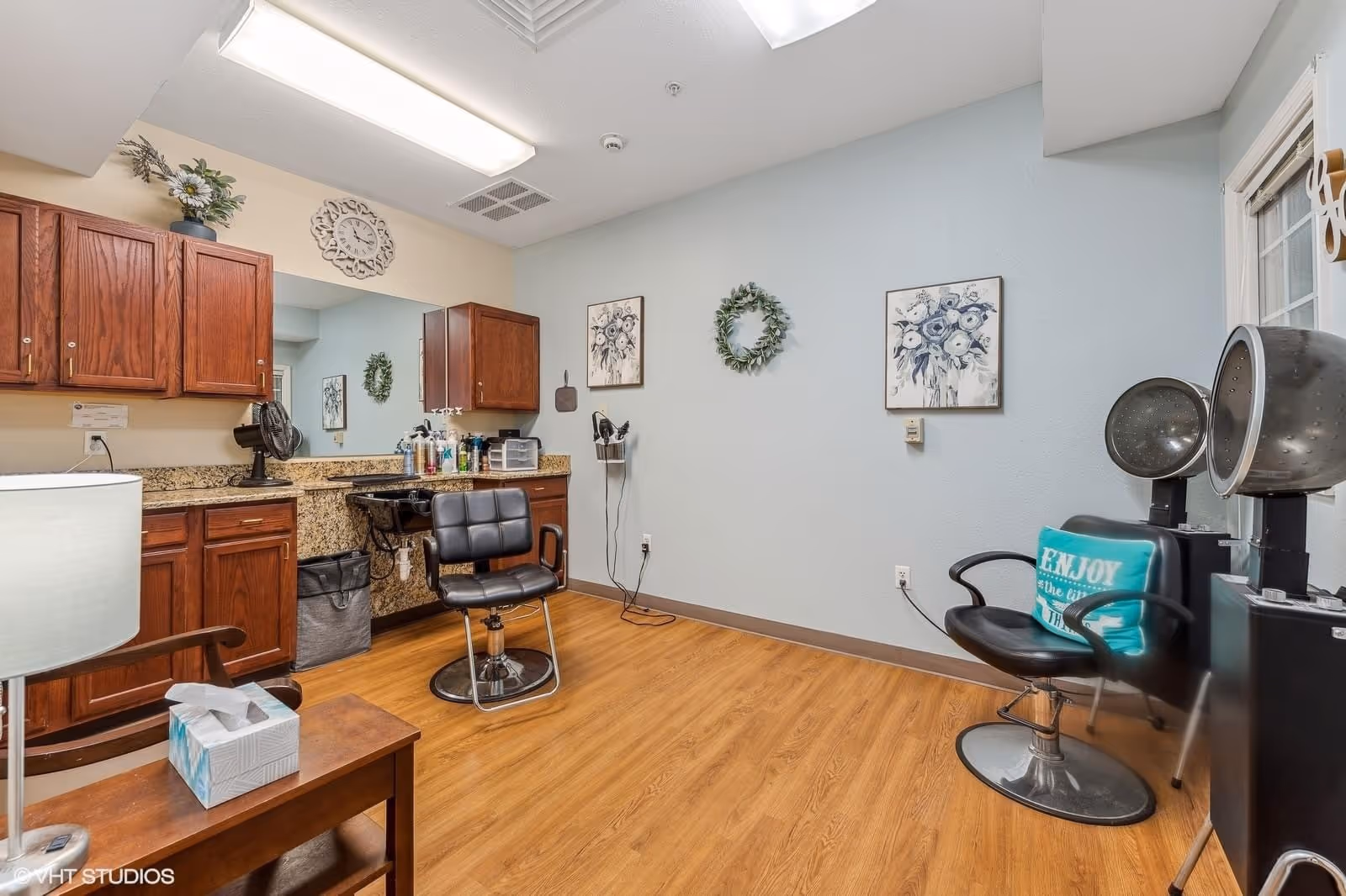 Interior salon room with styling chairs, hooded hair dryers, cabinets, and a sink in a senior living facility.