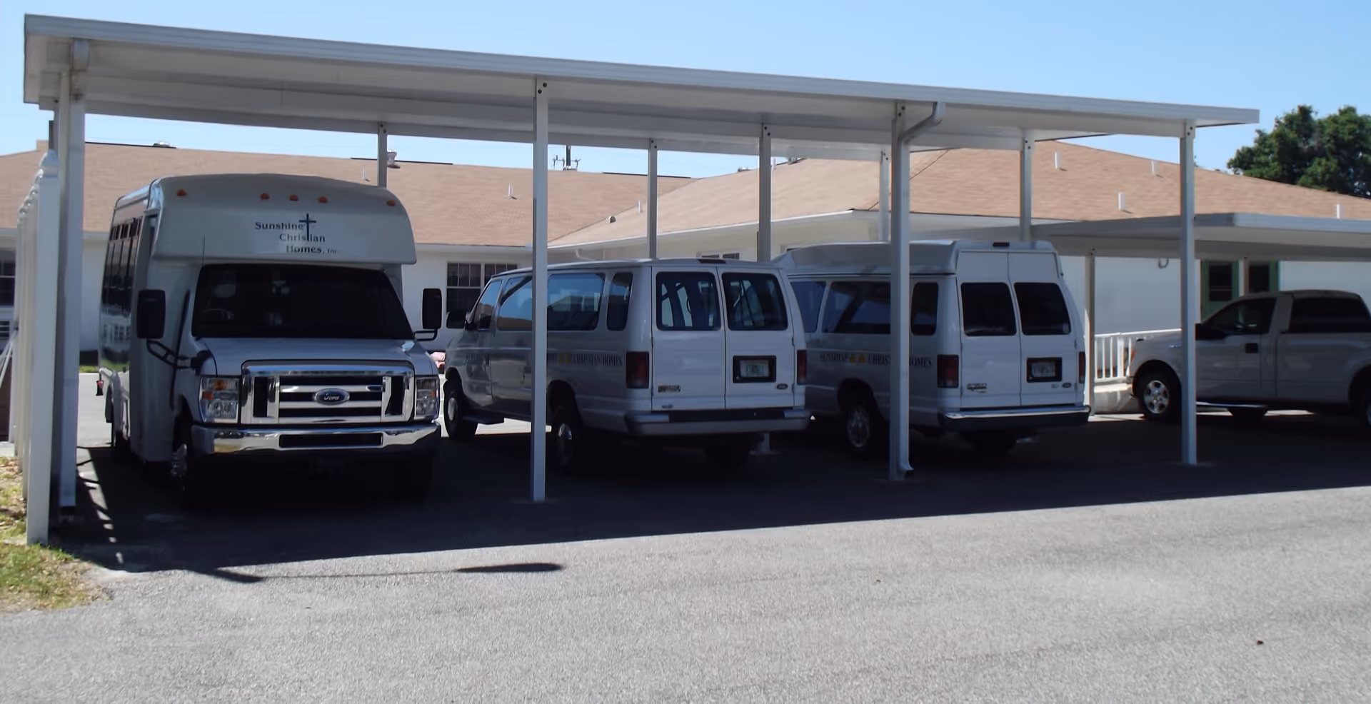 Several white vans and a shuttle bus parked under a covered carport in front of a single-story building.