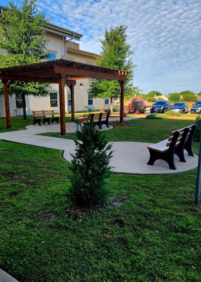 Outdoor seating area at Terry Lake Assisted Living featuring wooden benches on concrete paths surrounded by green grass and small trees, with a wooden pergola providing partial shade. In the background, there is a building with light-colored walls and several parked cars under a partly cloudy sky.