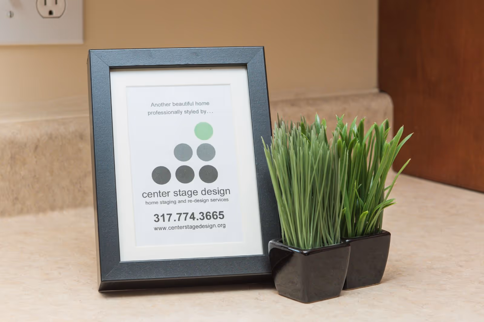 A black picture frame on a beige countertop displaying a card that reads 'Another beautiful home professionally styled by center stage design home staging and re-design services 317.774.3665 www.centerstagedesign.org' next to two small black pots with green artificial grass plants.