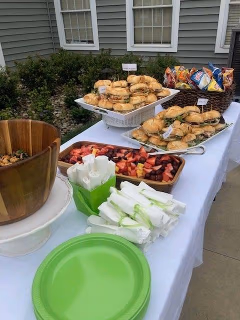 Outdoor buffet table set up with sandwiches, assorted chips in a basket, a tray of mixed fruit, wrapped utensils, and green disposable plates, placed in front of a building with gray siding and windows.