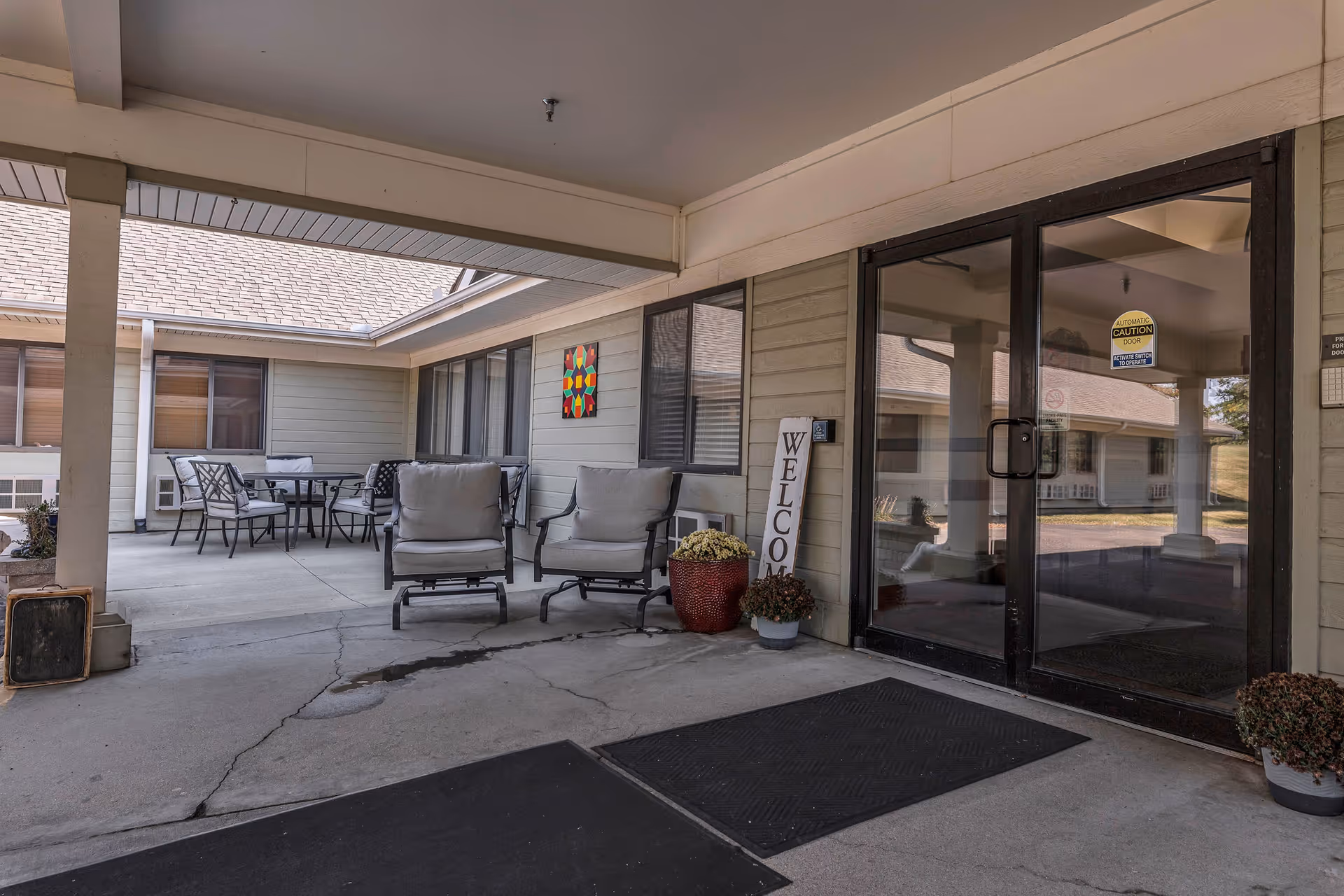 Covered entrance porch with chairs, a table, potted plants and glass double doors next to a 'Welcome' sign.