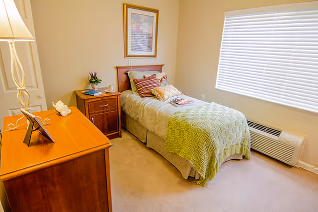 A tidy single bedroom with a bed, nightstand, dresser, lamp and a window with blinds.