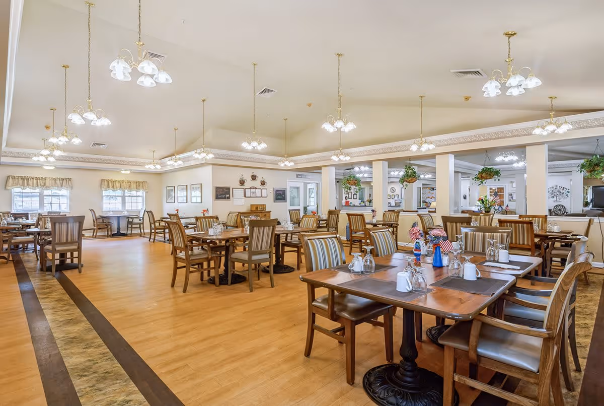 A spacious dining room with multiple wooden tables and chairs arranged neatly. The tables are set with placemats, glassware, and small decorative items. The room features light-colored walls, large windows with valances, hanging plants, and multiple ceiling light fixtures providing bright illumination.