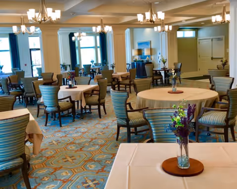 A spacious dining room with multiple round and square tables covered with beige tablecloths, each surrounded by striped upholstered chairs. The room features large windows with blue curtains, elegant chandeliers hanging from the ceiling, and a patterned carpet with blue and beige tones. A small vase with flowers is placed on the nearest table.