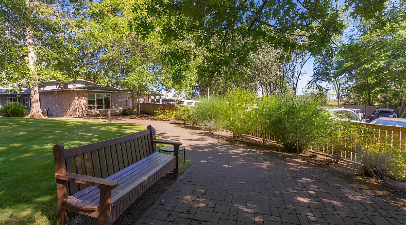 Wooden bench along a paved path in a shaded garden courtyard with lawn, trees, and a low building in the background.