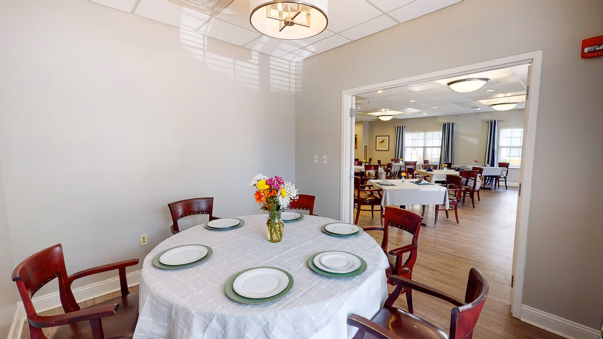 A dining area with a round table set with six plates and a vase of colorful flowers in the center. The room has wooden chairs and light-colored walls. Through an open doorway, a larger dining room with multiple tables and chairs is visible, featuring large windows with curtains and ceiling lights.