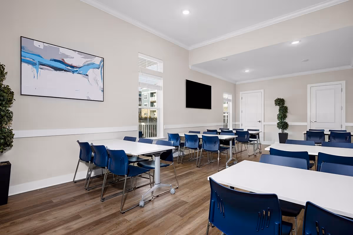 A bright and clean dining room with multiple white tables and blue chairs arranged neatly. The room has light-colored walls, wood flooring, two tall potted plants, a large abstract painting on one wall, and a flat-screen TV mounted on another wall. There are windows allowing natural light to enter the space.