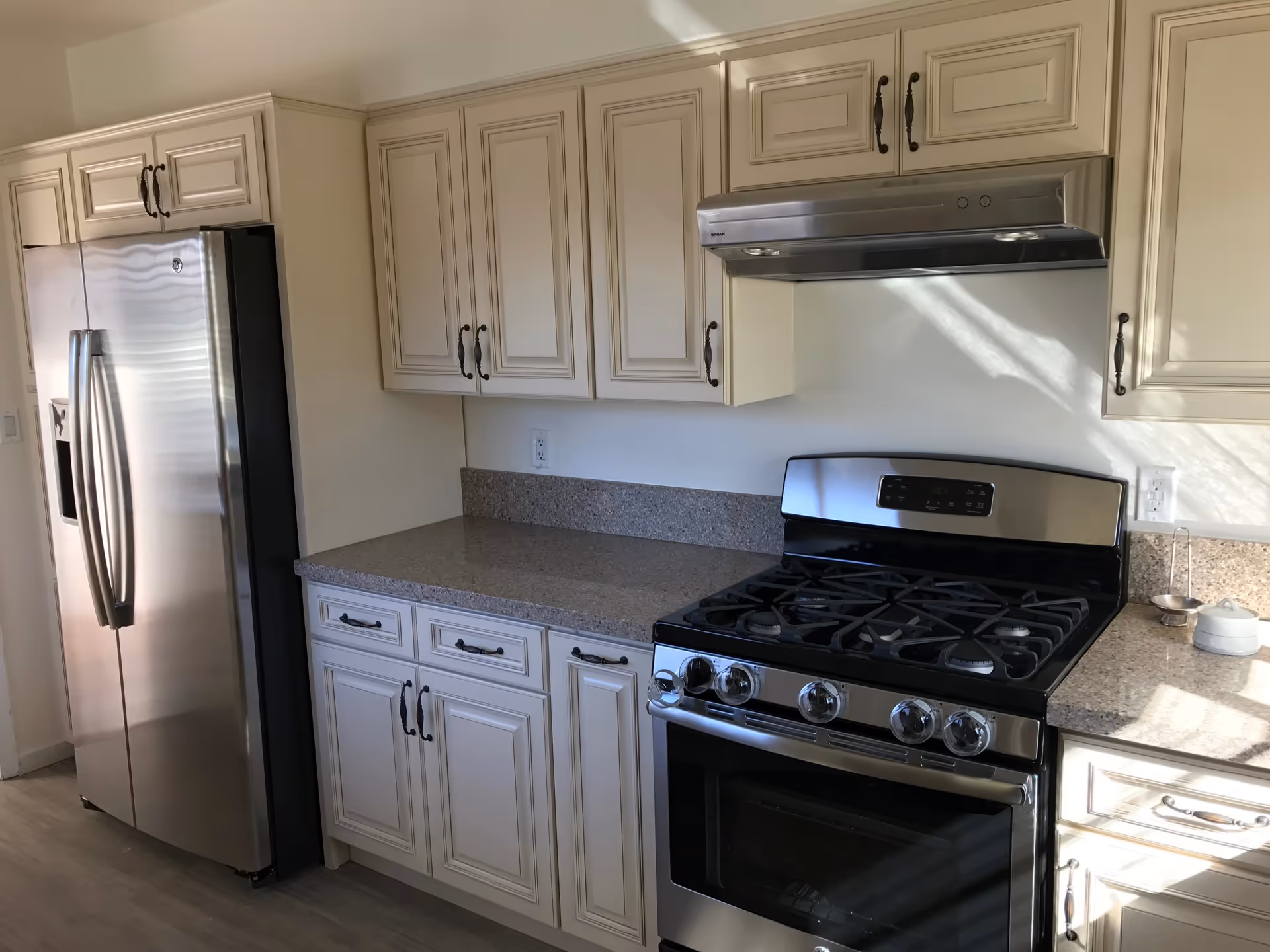 Modern kitchen with cream-colored cabinets, granite countertops, a stainless steel refrigerator, and a stainless steel gas stove with an overhead range hood.