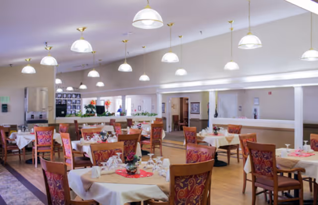 A spacious dining room with multiple tables covered in white tablecloths and set with glassware and floral centerpieces. The room features wooden chairs with patterned upholstery, hanging pendant lights, and a light-colored ceiling. The background shows a hallway and some framed pictures on the wall.