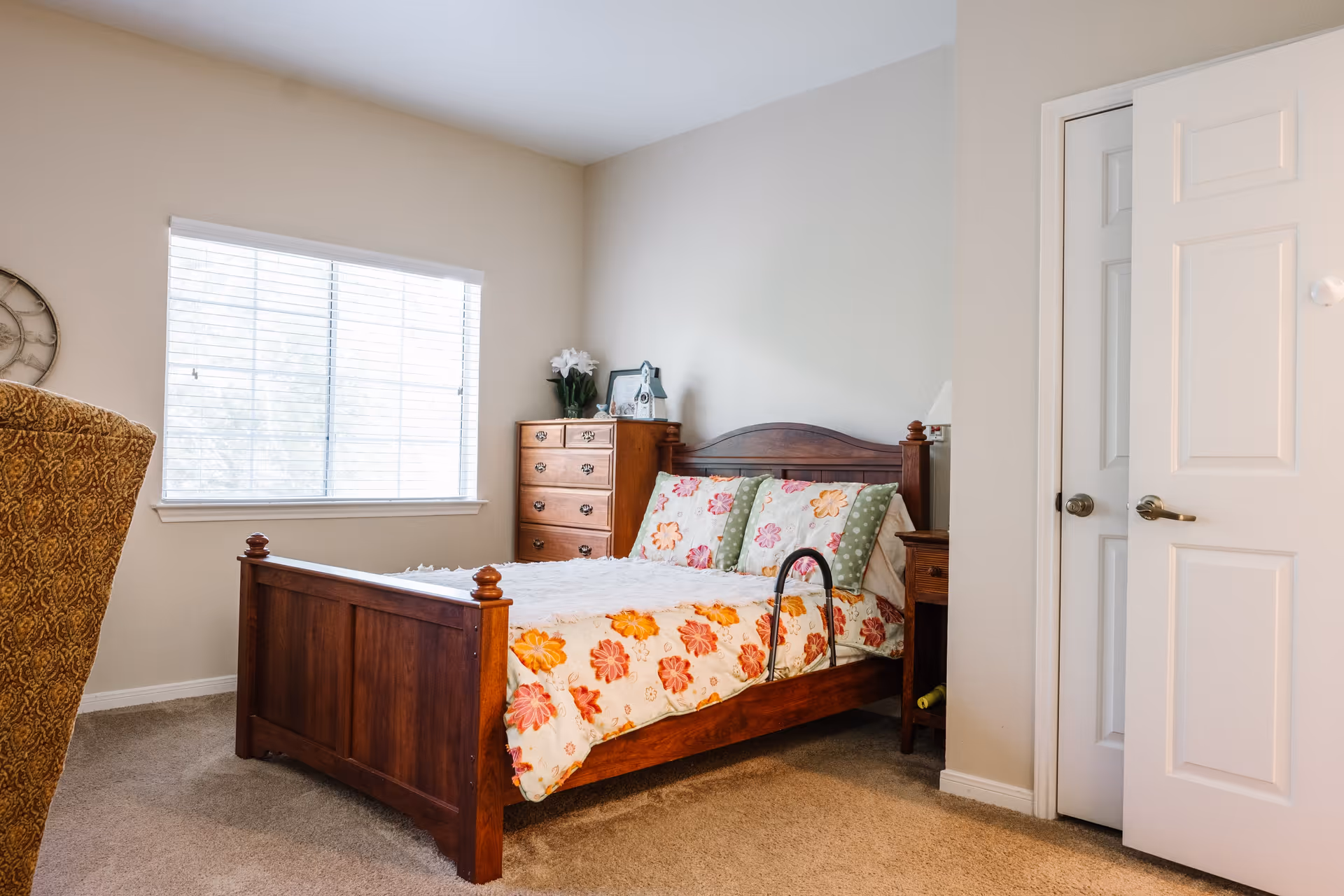 A cozy bedroom with a wooden bed frame featuring a floral bedspread and multiple pillows. There is a wooden dresser with decorative items and a window with blinds letting in natural light. A partially open white door is visible on the right side.