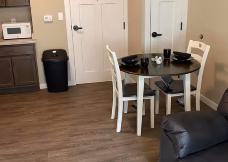 A small dining area with a round dark wood table set for two with black bowls and cups. Two white chairs with dark seats are placed around the table. To the left, there is a kitchen counter with a microwave on top and a black trash bin below. The floor is wood, and there are two white doors in the background. Part of a dark-colored sofa is visible on the right side.