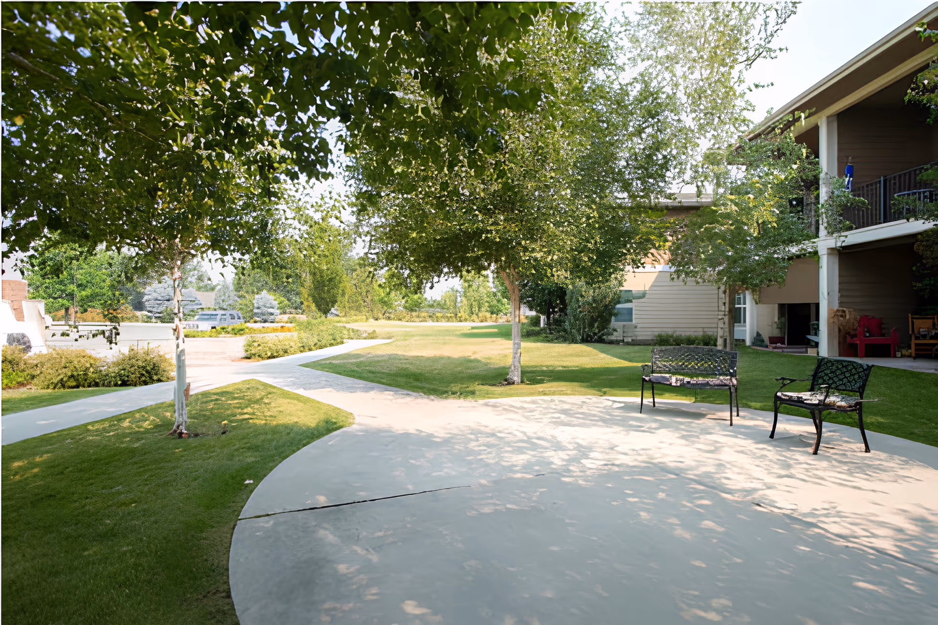 Outdoor area of Wenatchee Senior Living by Cogir featuring a concrete patio with two metal benches, surrounded by green grass, trees, and a building with balconies in the background.