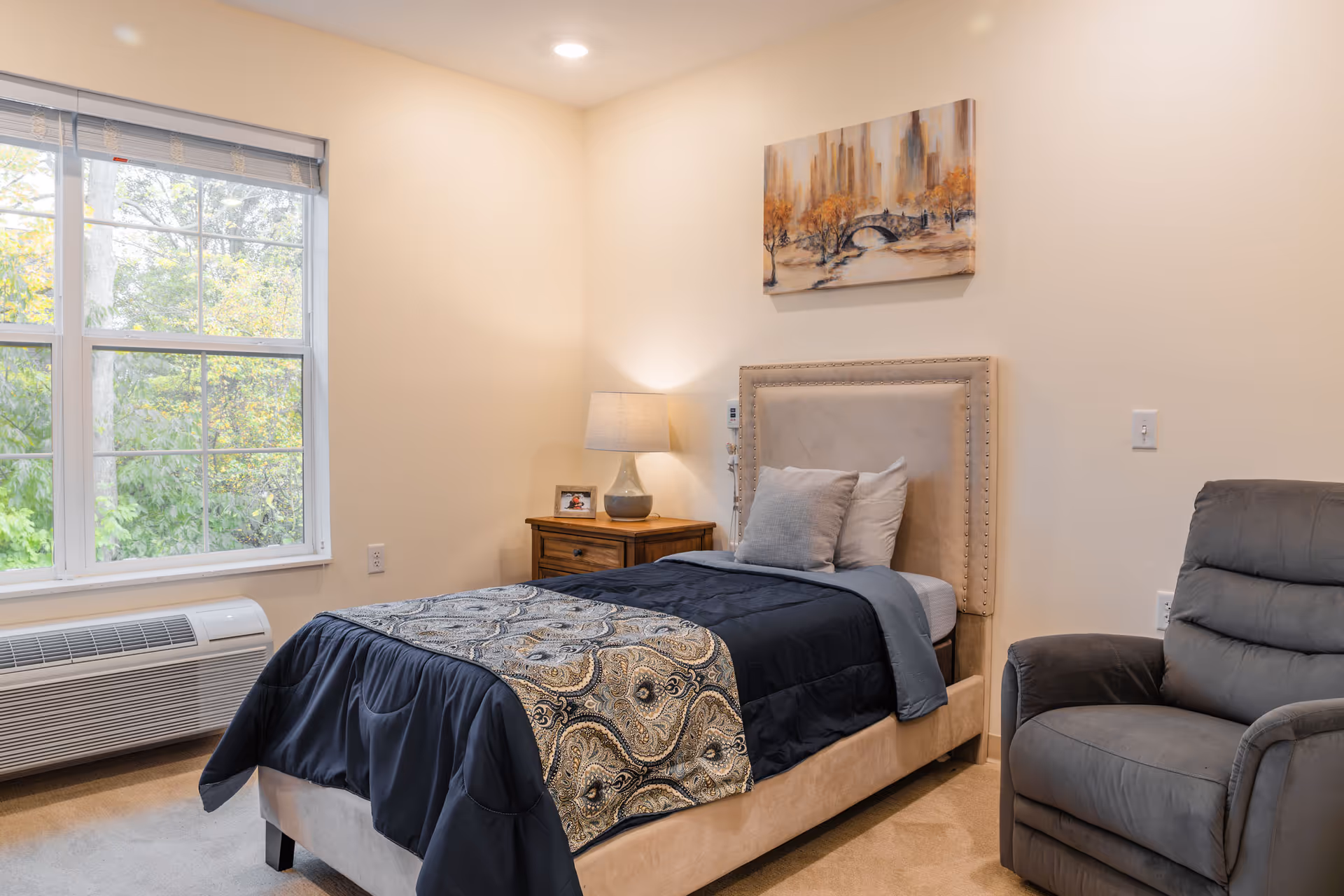 A cozy bedroom with a single bed featuring a beige upholstered headboard, dark blue and patterned bedding, and two pillows. Next to the bed is a wooden nightstand with a lamp and a small framed photo. A large window with a view of green trees lets in natural light. A comfortable gray recliner chair is positioned to the right of the bed. The walls are painted a soft cream color and a painting of a cityscape with autumn trees hangs above the bed.
